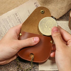 May include: A brown leather pouch with a gold-colored coin. The coin has the words "YOU ARE MY FAVORITE ADVENTURE" and a graphic. The pouch has a chain and a snap closure.