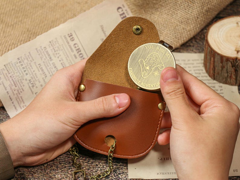 May include: A brown leather pouch with a gold-colored coin. The coin has the words "YOU ARE MY FAVORITE ADVENTURE" and a graphic. The pouch has a chain and a snap closure.
