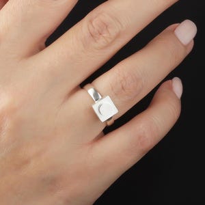 May include: A silver ring with a square face and a crescent moon design. The ring is displayed on a finger against a black backdrop. The ring's band has a smooth, simple design.