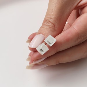 May include: A pair of square silver stud earrings, each with a small, circular accent. The earrings are held by a hand with manicured nails, set against a white background. The earrings are a minimalist design.