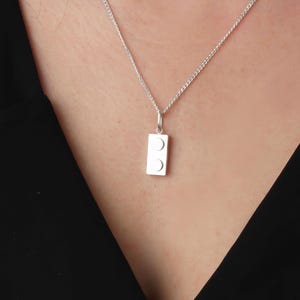 May include: A silver pendant necklace featuring a rectangular charm with two circular indentations. The pendant hangs from a delicate silver chain. The necklace is displayed against a dark background, highlighting the bright silver.
