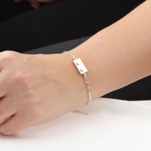 Può includere: Bracciale argentato con un ciondolo rettangolare con due dettagli circolari bianchi, che ricordano un mattoncino. Il bracciale ha un design a catena delicato, creando un accessorio moderno e giocoso.