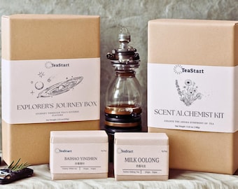 May include: Several tea boxes and a glass bottle with a decorative top are displayed. The boxes are tan with white labels featuring illustrations and text. The boxes are labeled "Explorer's Journey Box", "Scent Alchemist Kit", "Baihao Yinzhen", and "Milk Oolong".
