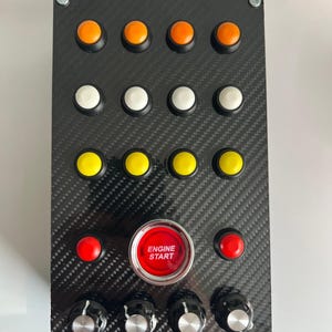 Könnte beinhalten: Eine schwarze Steuerungstafel mit Carbonfaser-Textur. Sie hat Reihen von orangefarbenen, weißen und gelben Knöpfen sowie einen großen roten "ENGINE START"-Knopf. Vier Drehknöpfe befinden sich unten.