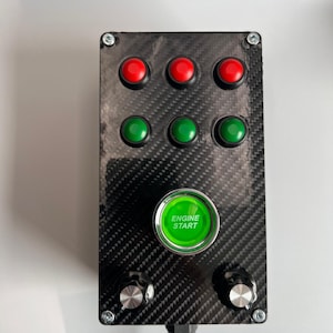 Op de afbeelding: Een zwart bedieningspaneel met een koolstofvezeltextuur. Het heeft zes ronde knoppen: drie rode en drie groene. Een grote groene knop in het midden leest "ENGINE START". Twee zilveren knoppen bevinden zich aan de onderkant. Een zwarte kabel komt van onderen.
