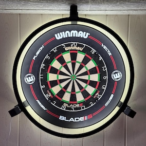 Puede incluir: Un tablero de dardos redondo negro con detalles rojos y blancos, con el logotipo de Winmau y el texto Blade 6. El tablero está montado sobre una superficie de madera y tiene una luz circundante.