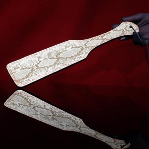 May include: A paddle with a snakeskin pattern in shades of white and tan, with red stitching around the edges. The paddle has a handle and a small hole for a cord. The image is reflected on a glossy surface.