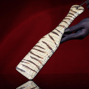 May include: A cream-colored paddle with brown tiger stripe pattern. The paddle has a handle with a hole for hanging. The paddle is held by a person wearing a black glove. The background is a dark red.