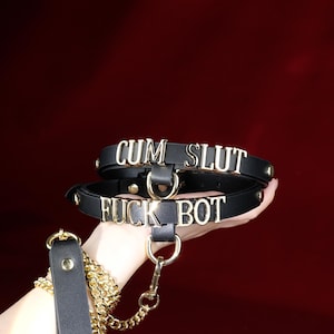 Op de afbeelding: Zwarte leren halsband met gouden letters met de tekst "CUM SLUT" en "FUCK BOT". De halsband heeft gouden hardware en een bijpassende riem met een gouden ketting. De achtergrond is donkerrood.