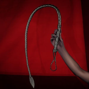 Peut inclure: Un fouet en cuir noir avec un manche tressé et une extrémité effilée. Le fouet est présenté sur un fond rouge, mettant en valeur sa texture et son design. La poignée comporte des nœuds décoratifs.