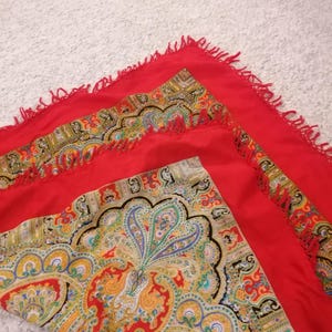 Può includere: Sciarpa quadrata rossa con un motivo paisley nei toni del giallo, verde, blu e rosso. La sciarpa ha un bordo con frange rosse. Il motivo paisley è concentrato negli angoli e lungo i bordi della sciarpa.