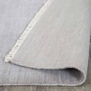 May include: A rolled-up, light gray rug with a textured surface. The rug's edge features a fringe detail. The underside of the rug is visible, showing a lighter shade of gray. The rug is made of a soft material.