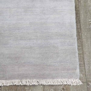 May include: A light gray rectangular rug with a fringe edge. The rug has a soft, textured appearance and is made of a woven material. The rug is displayed on a wooden floor.