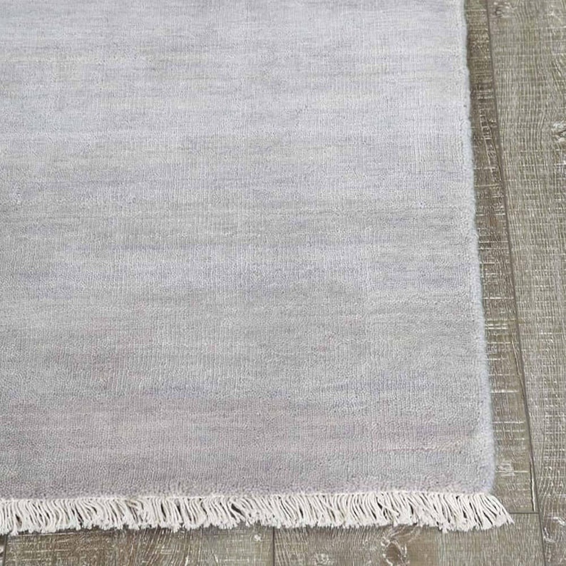 May include: A light gray rectangular rug with a fringe edge. The rug has a soft, textured appearance and is made of a woven material. The rug is displayed on a wooden floor.