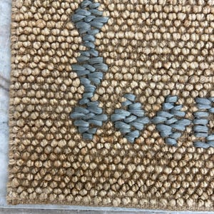 Puede incluir: Primer plano de una alfombra tejida con un fondo beige natural y un dise&ntilde;o geom&eacute;trico azul claro. La alfombra tiene una superficie texturizada y un borde. El dise&ntilde;o est&aacute; hecho de un material tejido.