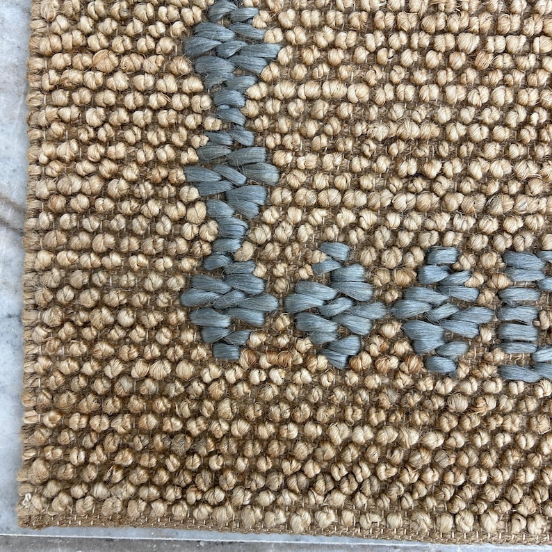 Puede incluir: Primer plano de una alfombra tejida con un fondo beige natural y un dise&ntilde;o geom&eacute;trico azul claro. La alfombra tiene una superficie texturizada y un borde. El dise&ntilde;o est&aacute; hecho de un material tejido.