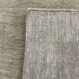 May include: A close-up of a beige rug with a textured surface. The rug's corner is visible, showcasing a detailed edge. The neutral color and woven texture suggest a home decor item.