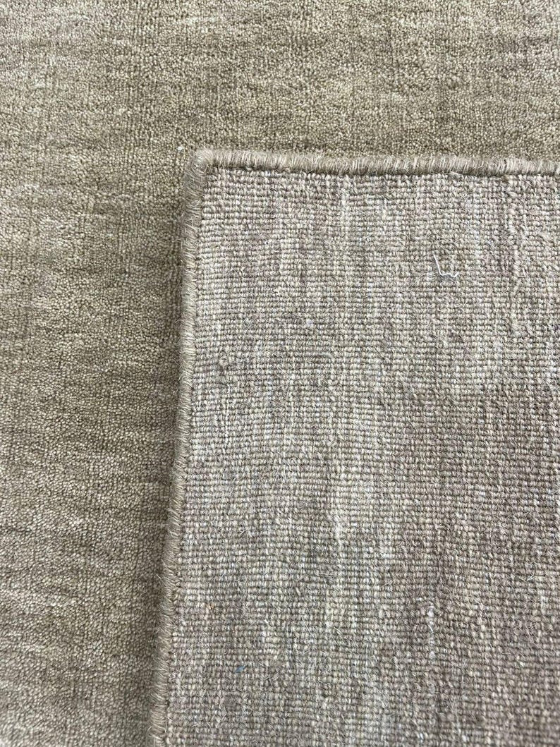 May include: A close-up of a beige rug with a textured surface. The rug's corner is visible, showcasing a detailed edge. The neutral color and woven texture suggest a home decor item.