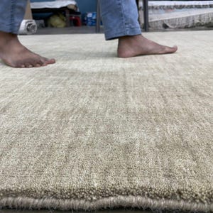 May include: A close-up shot of a beige rug with a textured surface. The rug has a soft, plush appearance and a subtle pattern. The image shows the edge of the rug and a person's feet and legs in the background.