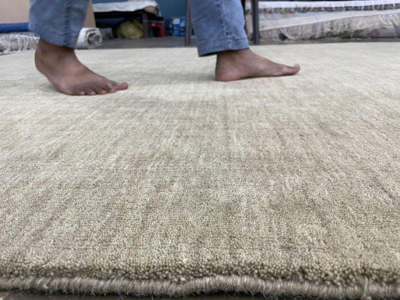 May include: A close-up shot of a beige rug with a textured surface. The rug has a soft, plush appearance and a subtle pattern. The image shows the edge of the rug and a person's feet and legs in the background.