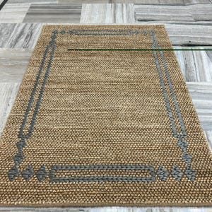 Puede incluir: Una alfombra rectangular de yute tejido, de color beige natural. Presenta un borde gris tejido, creando un dise&ntilde;o enmarcado. La textura rugosa de la alfombra sugiere durabilidad, adecuada para zonas de alto tr&aacute;nsito.