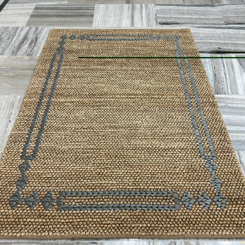 Puede incluir: Una alfombra rectangular de yute tejido, de color beige natural. Presenta un borde gris tejido, creando un dise&ntilde;o enmarcado. La textura rugosa de la alfombra sugiere durabilidad, adecuada para zonas de alto tr&aacute;nsito.