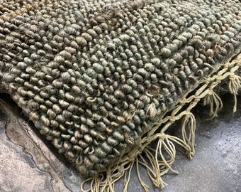 May include: Close-up of a textured, hand-woven rug. The rug features a mix of green, brown, and beige tones, with a dense, looped pile. The edge of the rug has a fringe detail in a lighter beige colour, adding a rustic touch.