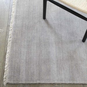 May include: A light gray rectangular rug with a fringed edge. The rug has a textured surface and is partially covered by a black chair leg and a portion of a woven chair seat. The rug is placed on a wooden floor.