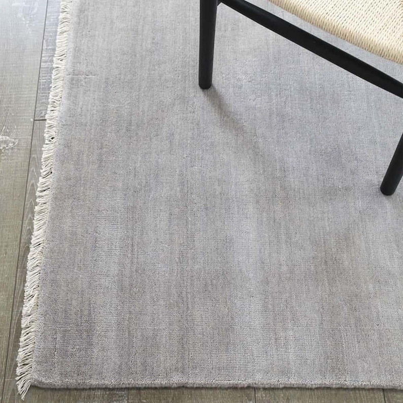 May include: A light gray rectangular rug with a fringed edge. The rug has a textured surface and is partially covered by a black chair leg and a portion of a woven chair seat. The rug is placed on a wooden floor.