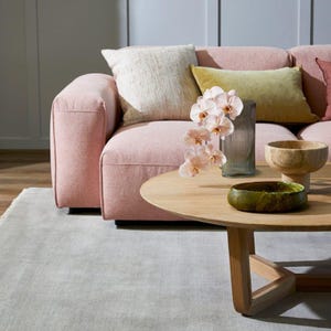 May include: A modern living room scene featuring a pink sofa with decorative pillows in cream and olive green. A wooden coffee table displays a vase with orchids, a wooden bowl, and a green decorative bowl. A light gray rug is in the foreground.