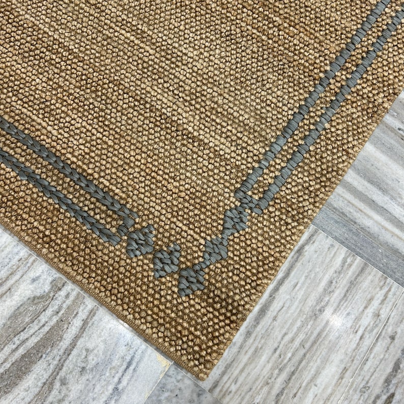 Puede incluir: Primer plano de una alfombra rectangular de yute tejido de color beige natural. La alfombra presenta un borde de detalles tejidos grises a lo largo de los bordes, creando un patr&oacute;n decorativo. La alfombra est&aacute; colocada sobre un suelo de m&aacute;rmol.