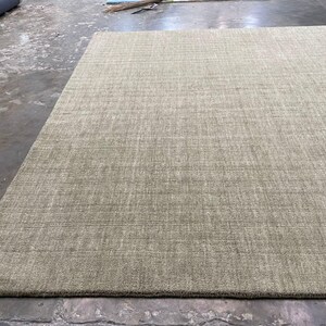 May include: A large, rectangular area rug in a neutral beige color. The rug has a textured surface with subtle linear patterns, suggesting a woven construction. The rug is placed on a concrete floor.