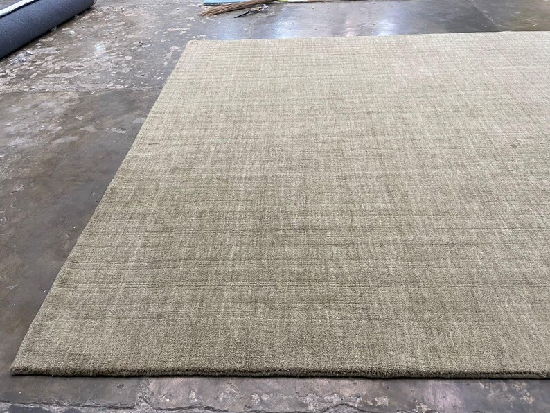 May include: A large, rectangular area rug in a neutral beige color. The rug has a textured surface with subtle linear patterns, suggesting a woven construction. The rug is placed on a concrete floor.