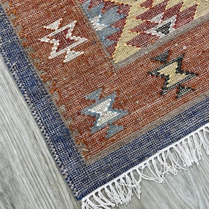 Könnte beinhalten: Nahaufnahme eines gewebten Teppichs mit geometrischem Muster. Der Teppich hat einen dunkelblauen Rand und ein zentrales Design in Rost-, Beige- und Grautönen. Weiße Fransen akzentuieren den Rand. Das Design des Teppichs umfasst sternartige Formen.