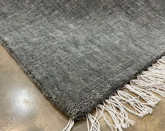 May include: A rectangular area rug featuring a dense, textured pile in a solid charcoal grey colour. The rug is finished with a row of off-white, knotted fringe tassels along the edge, providing a contrasting detail to the deep grey surface.