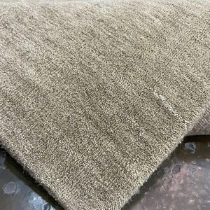May include: Close-up of a beige rug with a textured, looped pile. The rug's corner is folded over, revealing the underside. The neutral color and dense texture suggest a durable and versatile floor covering suitable for various interior styles.