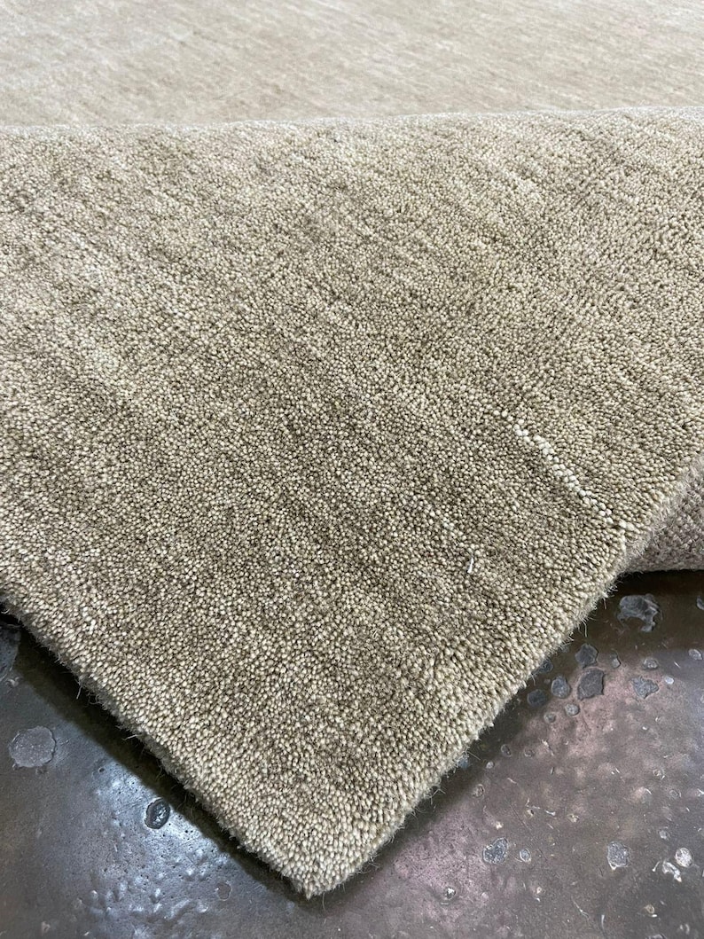 May include: Close-up of a beige rug with a textured, looped pile. The rug's corner is folded over, revealing the underside. The neutral color and dense texture suggest a durable and versatile floor covering suitable for various interior styles.