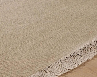 May include: A beige rectangular rug with a textured surface and a fringe border. The rug is made of a woven material and is placed on a wooden floor. The rug's neutral color makes it suitable for various interior design styles.