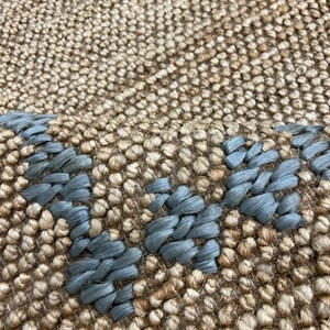 Puede incluir: Primer plano de una alfombra tejida con un fondo beige natural y un dise&ntilde;o geom&eacute;trico azul. La alfombra tiene una superficie texturizada y una paleta de colores neutros, adecuada para varios estilos de interior.