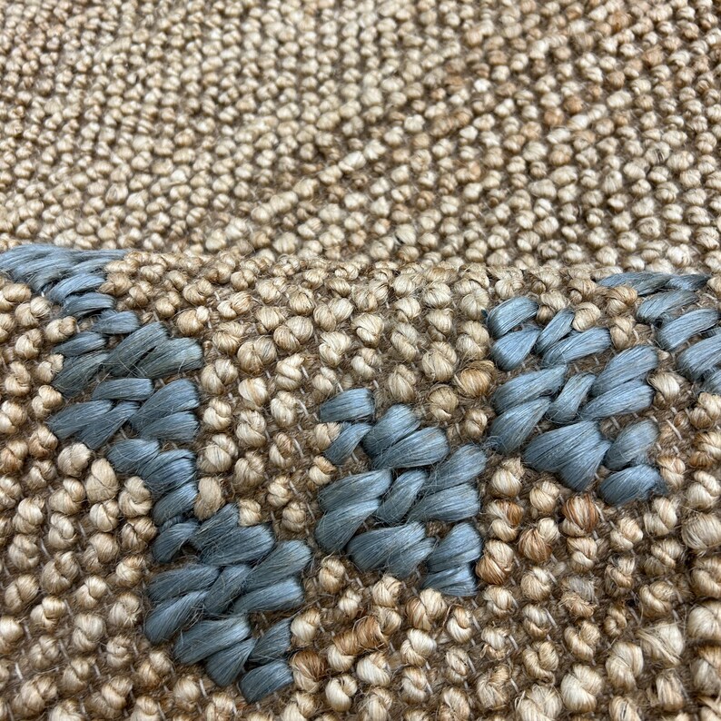 Puede incluir: Primer plano de una alfombra tejida con un fondo beige natural y un dise&ntilde;o geom&eacute;trico azul. La alfombra tiene una superficie texturizada y una paleta de colores neutros, adecuada para varios estilos de interior.