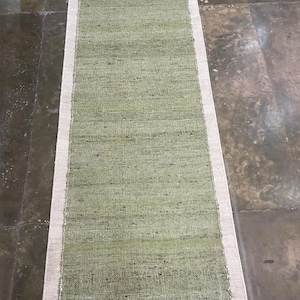 Può includere: Un tappeto rettangolare verde chiaro con un bordo color crema. Il tappeto ha una trama tessuta ed è probabilmente realizzato con fibre naturali. È progettato per un corridoio o un ingresso, misurando circa 150 cm di lunghezza.