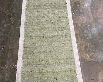 May include: A rectangular, pale green rug with a cream border. The rug has a woven texture, possibly made from natural fibres. The rug is suitable for a hallway or entryway, measuring approximately 150 cm in length.