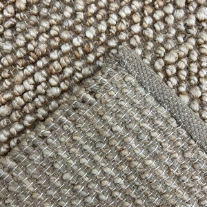 Puede incluir: Primer plano de una alfombra de fibra natural con una superficie texturizada y nudosa. La alfombra presenta un color marr&oacute;n claro con un borde tejido y costuras blancas. La esquina de la alfombra est&aacute; doblada, revelando la parte inferior.