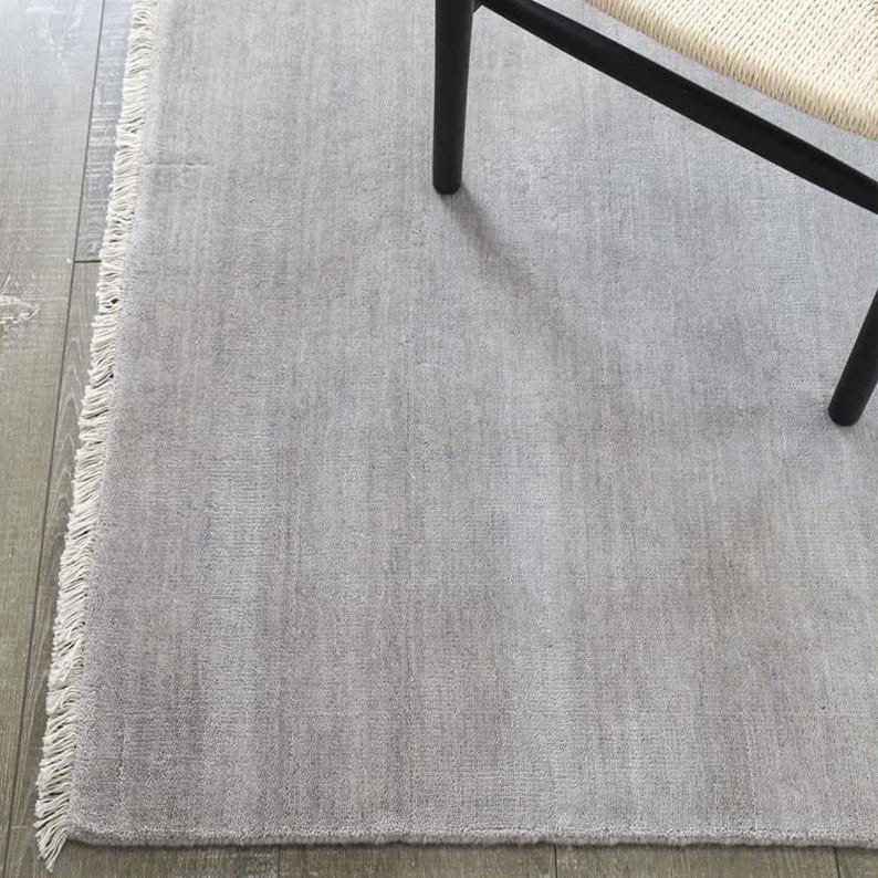 May include: A light gray rectangular rug with a fringed edge. The rug has a textured surface and is partially covered by a black chair leg and a portion of a woven chair seat. The rug is placed on a wooden floor.