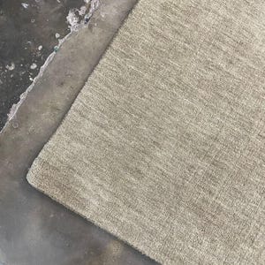May include: A rectangular, beige rug with a textured surface. The rug is positioned diagonally against a dark, mottled surface. The rug's fibers appear soft and plush, suggesting a comfortable texture.