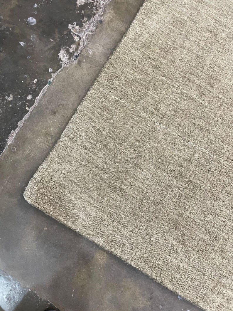 May include: A rectangular, beige rug with a textured surface. The rug is positioned diagonally against a dark, mottled surface. The rug's fibers appear soft and plush, suggesting a comfortable texture.