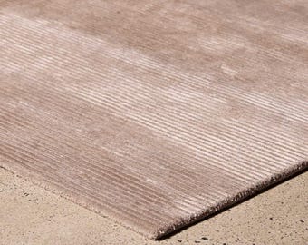 May include: A rectangular area rug in a muted taupe color featuring a consistent, fine-ribbed linear texture across its entire surface. The rug has a clean, straight edge and a uniform, low-pile appearance with subtle light reflections.