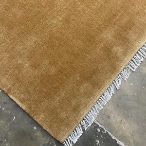 Könnte beinhalten: Ein einfarbiger, senfgelber Teppich mit kurzem, dichtem Flor. Der rechteckige Teppich hat eine gerade Kante, die mit einer Reihe weißer, gedrehter Fransen versehen ist, was für ein sauberes und gleichmäßiges Erscheinungsbild sorgt.