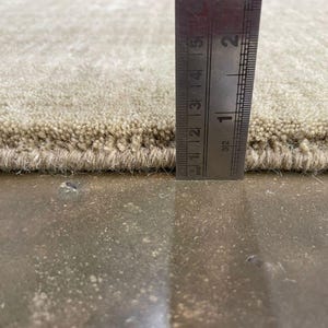 May include: Close-up of a beige rug with a textured surface. A metal ruler is placed vertically against the rug, showing a thickness of approximately 0.5 inches. The rug's edge is visible, revealing its construction.