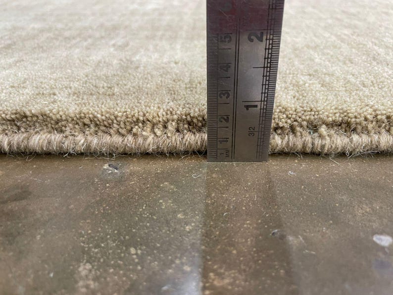 May include: Close-up of a beige rug with a textured surface. A metal ruler is placed vertically against the rug, showing a thickness of approximately 0.5 inches. The rug's edge is visible, revealing its construction.